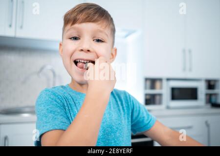 ragazzo felice mangiare o mordere dolci al cioccolato o caramelle in moderna cucina casalinga Foto Stock