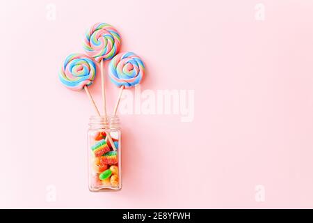 I lollipop colorati roteano su bastoni in una bottiglia di vetro. Caramelle multicolore a spirale su sfondo rosa, vista dall'alto Foto Stock