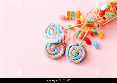 I lollipop colorati roteano su bastoni in una bottiglia di vetro. Caramelle multicolore a spirale su sfondo rosa, vista dall'alto Foto Stock