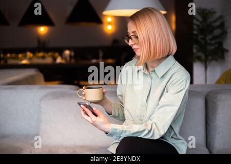 Una ragazza si siede in un caffè con un telefono in la sua mano e tiene una tazza di caffè Foto Stock