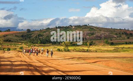 Sendrisoa, Madagascar - 28 aprile 2019: Gruppo di uomini, donne e bambini malgasci sconosciuti che camminano a casa dopo aver lavorato nei campi, sulla strada della polvere rossa, gr Foto Stock