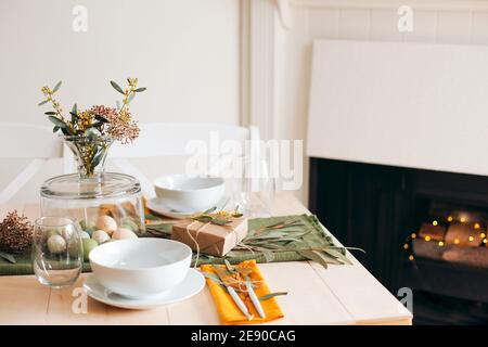 Tavolo di legno bianco con piatto e ciotola bianchi, uova di Pasqua di colore pastello, fiori freschi del giardino e carta marrone da regalo, arredamento del soggiorno Foto Stock
