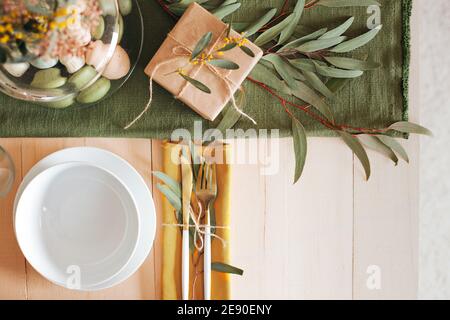 Tavolo di legno bianco con piatto e ciotola bianchi, uova di Pasqua color pastello, fiori freschi da giardino e carta marrone in confezione regalo, runner da tavolo verde Foto Stock