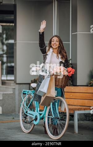 Bella giovane donna sorridente che cammina in bicicletta, dopo aver fatto shopping e ondulato qualcuno di mano, sulla strada della città. Bellezza, gesto di saluto e stile di vita Foto Stock