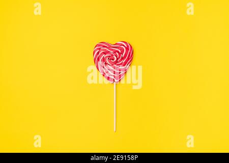 Caramella lollipop rossa a forma di cuore isolata su uno sfondo vuoto colorato giallo. Simbolo d'amore per le donne felici, San Valentino, compleanno saluto cartolina romantica. Disposizione piatta, vista dall'alto, spazio per la copia Foto Stock