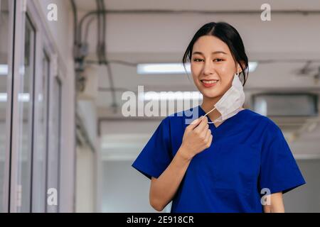 Giovane bella infermiera medica asiatica in uniforme scrub all'ospedale preparato a indossare una maschera facciale. Medica professionale sul posto di lavoro durante la pandemia di Covid19. Foto Stock