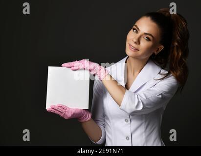 Giovane bruna sorridente donna medico terapista infermiera in uniforme bianca e guanti di protezione che tengono fogli di carta bianchi Foto Stock