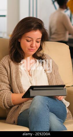 Donna manager che cerca sul tablet seduto nella zona relax sul comodo divano sorridente, puntando sul blocco note sorridendo mentre diversi colleghi lavorano in background. Collaboratori che pianificano un progetto finanziario Foto Stock