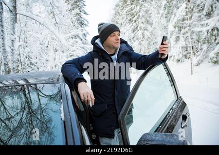 Uomo in abiti caldi invernali in piedi in auto, utilizzando il telefono. Neve strada di campagna invernale, auto coperta di ghiaccio, bella foresta sotto la neve. Foto Stock