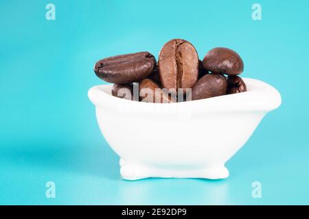 Primo piano di una piccola vasca da bagno bianca piena di chicchi di caffè tostati su uno sfondo azzurro. Concetto creativo di routine mattutina. Studio shot, foc. Selettivo Foto Stock