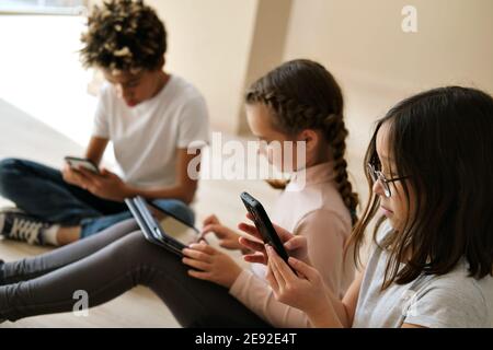 Tre bambini africani, caucasici e coreani multirazziali si siedono all'interno utilizzando i gadget wireless. Uso eccessivo di smartphone tecnologici moderni, dispositivi digitali o Foto Stock