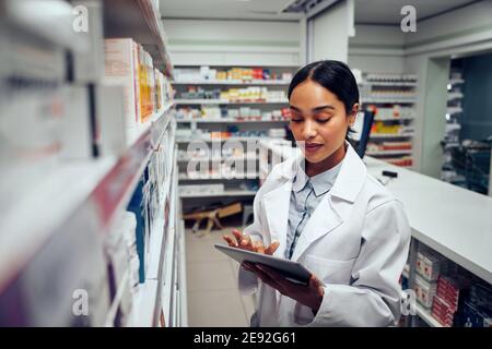 Giovane donna farmacista controllo inventario dei medicinali in farmacia usando tablet digitale con camice da laboratorio in piedi dietro il bancone Foto Stock