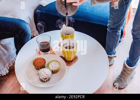 Donna che serve il tè con un bollitore da stiro in tazze su un piccolo tavolo con cupcake. Interni domestici Foto Stock