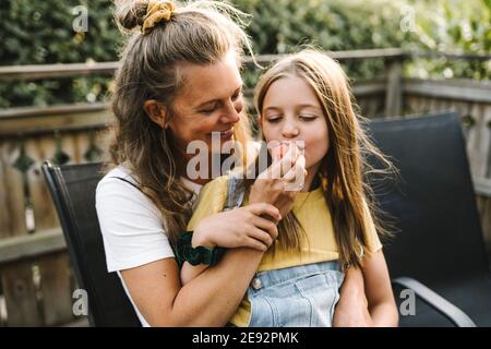 Madre sorridente che alimenta il cocomero alla figlia mentre si siede nel balcone Foto Stock