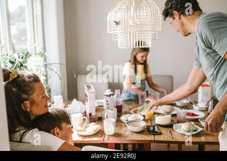 Madre e figlio si abbracciano mentre padre e figlia si levano in piedi vicino al tavolo da pranzo Foto Stock