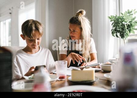 La madre insegna mentre il figlio usa lo smartphone a casa Foto Stock