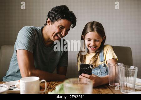 Sorridente padre e figlia utilizzando il telefono cellulare al tavolo da pranzo Foto Stock