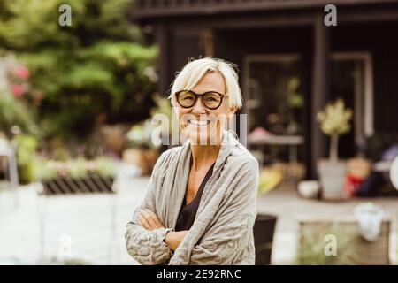 Donna matura sorridente con braccia incrociate in cortile posteriore Foto Stock