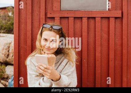 Donna sorridente con la mano sul mento che tiene lo smartphone contro cottage Foto Stock