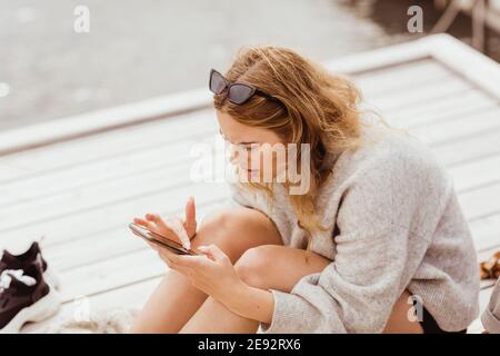 Donna bionda che usa lo smartphone mentre si siede sul molo Foto Stock