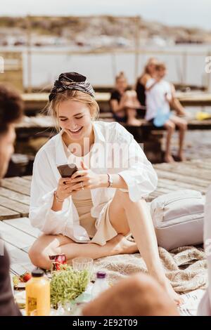 Donna sorridente che usa lo smartphone seduto da un amico sul molo durante il picnic Foto Stock