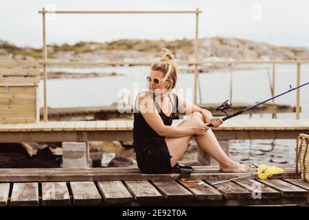 Donna matura con canna da pesca che guarda sulla spalla mentre si siede sul molo al porto Foto Stock