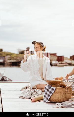 Donna sorridente che prende selfie seduto sul molo contro cielo durante picnic Foto Stock