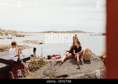 Madre e figlia che si siedono sulla roccia mentre fratello si siede con canna da pesca durante la giornata di sole Foto Stock