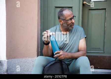 Uomo anziano con bottiglia che guarda via mentre si siede contro la porta Foto Stock