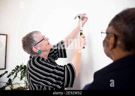 Donna sorridente che batte il chiodo sulla parete mentre si sta in piedi da senior uomo a casa Foto Stock