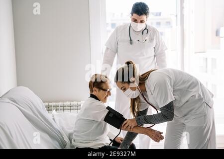 Medici in maschere mediche utilizzando tonometro e la misurazione della pressione sanguigna di anziana paziente femminile seduta su divano a casa Foto Stock