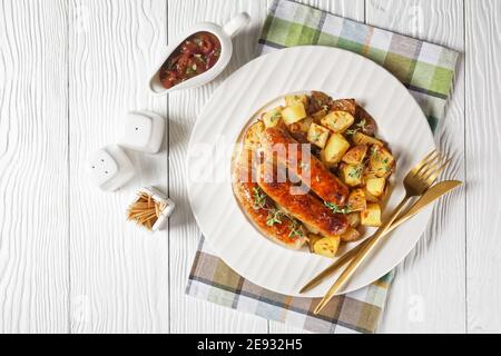 salsicce e patate arrostite su un piatto bianco con sugo di cipolla in una caraffa su un tavolo di legno testurizzato bianco, piatto Foto Stock