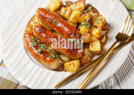 bangers e patate arrostite su un piatto bianco con cipolla salate in una caraffa su un tavolo di legno con trama bianca Foto Stock