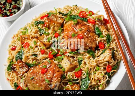 Cucina cinese: Tagliatelle con cosce di pollo dolci e acide, tofu al forno, kale selvatico, peperoncino rosso, cipolla verde, su un piatto bianco con bastoncini Foto Stock