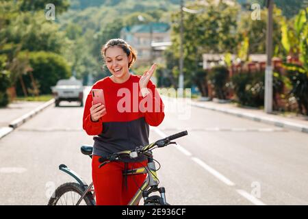 Attività. Una giovane bella donna è seduta su una bicicletta, e felicemente agita la mano mentre fa una videochiamata. All'aperto. Concetto di tecnologia moderna Foto Stock
