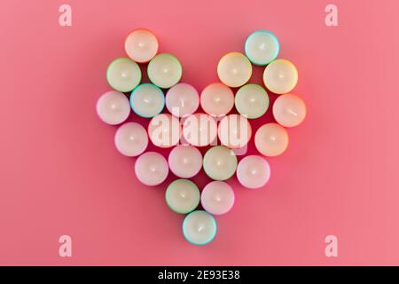 Vista dall'alto di file di candele inutilizzate multicolore di cera da tè disposte a forma di cuore su sfondo rosa. Fila di nuove candele d'acqua come backgr Foto Stock