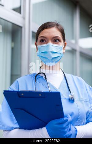Giovane donna caucasica UK NHS medico GP guardando a distanza Con paura, preoccupazione, ansia negli occhi, indossare blu ICU uniforme maschera viso, speranza e. fede in overc Foto Stock