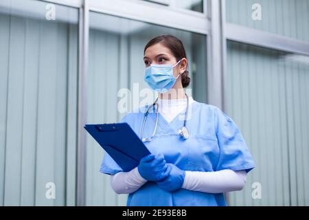 Donna UK NHS ICU medico lavoratore, donna medico tenendo appunti indossando Maschera protettiva blu PPE, medica di emergenza anteriore, COVID-19 pande Foto Stock