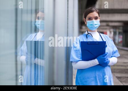 Giovane donna NHS UK EMS medico di fronte alla sanità Struttura ICU, indossare una maschera protettiva per il viso che tiene il controllo medico delle condizioni del paziente forma, riflessione i Foto Stock