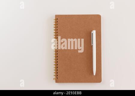 Quaderno marrone chiuso a spirale e penna bianca. Layout e disposizione piatta con posizione per il testo isolato su sfondo bianco. Ritorno a scuola. Vista dall'alto, spazio di copia Foto Stock