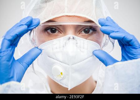 Femmina NHS medico, infermiere o laboratorio tecnico virologia scienziato in PPE, mettere sugli occhiali di protezione, indossare guanti in lattice blu e il viso bianco maschera, clos Foto Stock