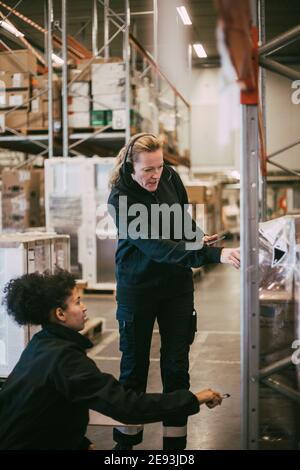 Donna d'affari che indossa le cuffie facendo il controllo di qualità dei pacchetti con la donna collega in magazzino Foto Stock