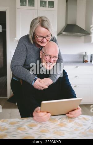 Una coppia sorridente che si è abbracciata e ha una chat video sul tablet a. tavolo da cucina Foto Stock
