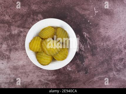 Vista dall'alto di una piccola ciotola bianca piena di fette di cetriolo di hamburger sfalsate su un tavolo di maroon illuminato con illuminazione naturale. Foto Stock