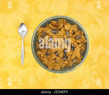 Vista dall'alto di una ciotola piena di fiocchi di crusca e uvetta per la colazione con un cucchiaio a lato su una tovaglia floreale gialla. Foto Stock