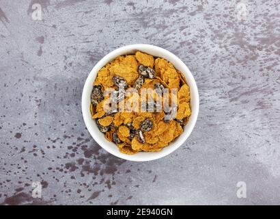 Vista dall'alto di una ciotola bianca piena di fiocchi di crusca e uvetta per la colazione, cereali su sfondo grigio. Foto Stock