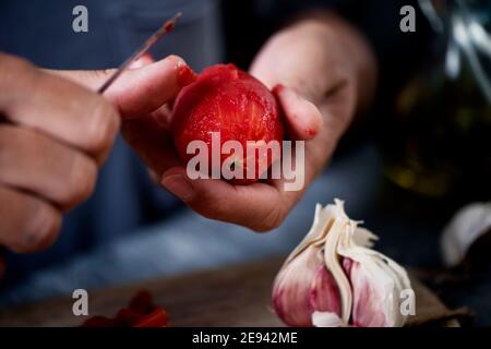 primo piano di un giovane caucasico, indossando una t-shirt grigia, sbucciando un pomodoro scottato su un tavolo da cucina o piano accanto ad una testa di aglio Foto Stock