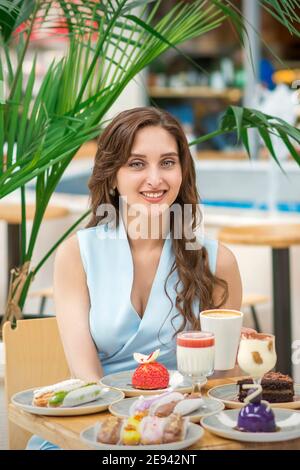 Una bella giovane caucasica seduta al tavolo con torte e una tazza di caffè nella caffetteria all'aperto Foto Stock