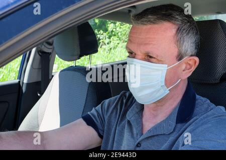Un adulto di mezza età si siede dietro la ruota della sua auto indossando una maschera medica. Influenza, coronavirus, quarantena, concetto di autoisolamento. Protectio Foto Stock
