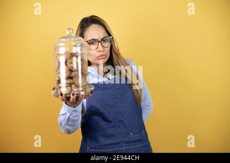 Giovane donna ispanica che indossa un panettiere uniforme tenendo e mostrando un vasetto di biscotti Foto Stock